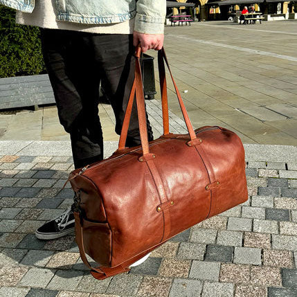 Person holding a brown leather duffel bag in an outdoor setting with historical architecture.