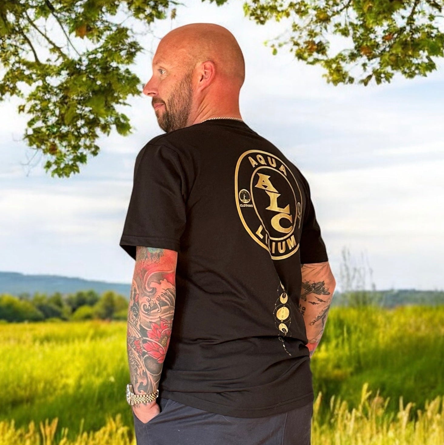 Man wearing a black t-shirt with a logo standing in a grassy field rokkatei-eshop.mom