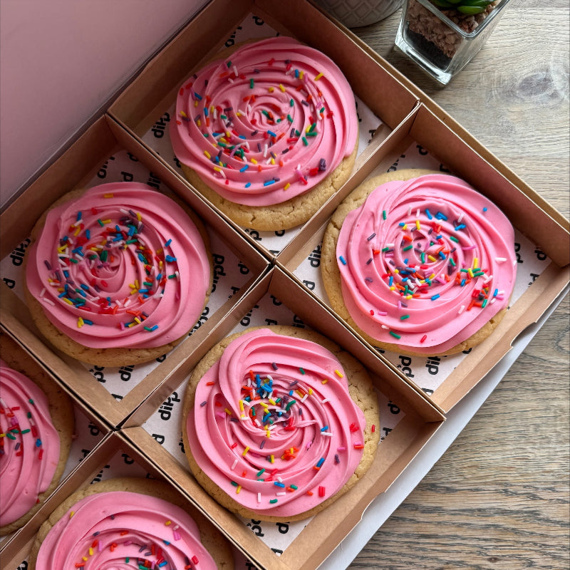 Box of cookies with pink frosting and sprinkles on a wooden surface on Sostter