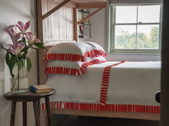Bed with red and white striped bedding in a room with a window and small table with flowers and books. Brand Sukun on Sostter
