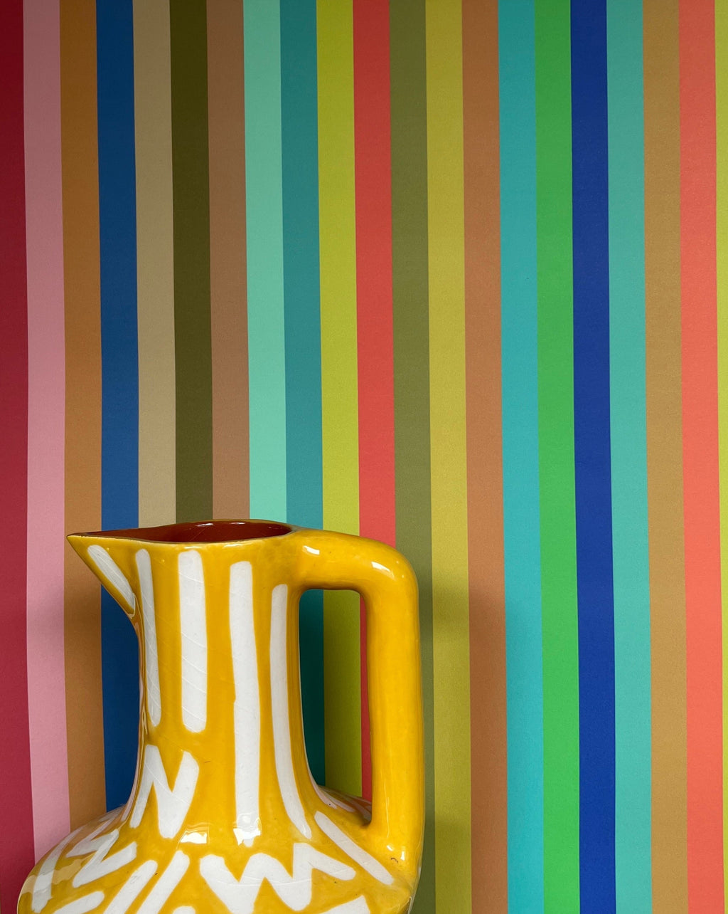 Yellow pitcher with white patterns against a multicolored striped wall bySharon Jane Studio at Sostter