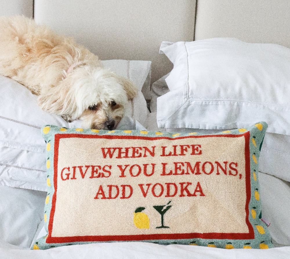 Dog lying on a bed with a decorative pillow that says 'When life gives you lemons, add vodka'. From The Pillow Drop at Sostter