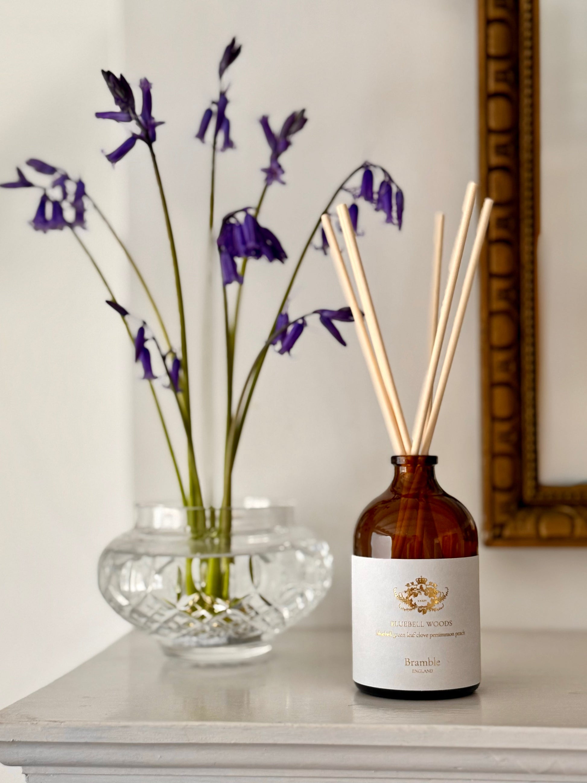 Diffuser bottle with reeds and a vase of purple flowers on a surface.