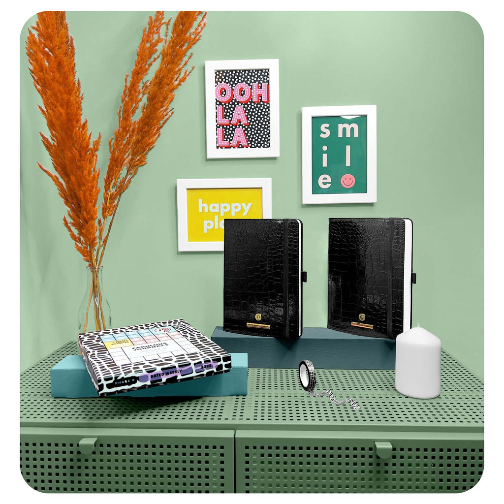 Decorative setup with black folders, a vase with orange plants, and framed pictures on a green surface on Sostter