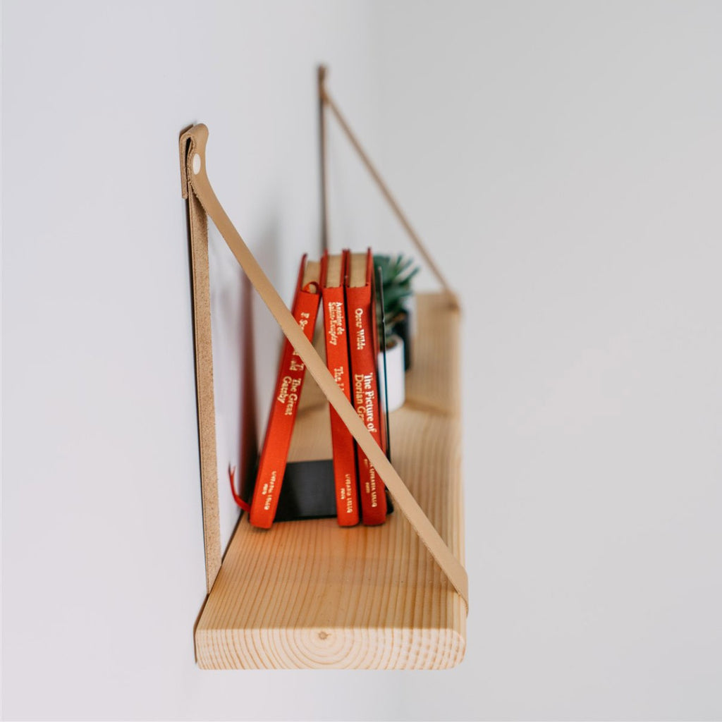 a floating pine shelf with camel leather straps in a nursery setting, holding soft baby clothing and a small plush toy