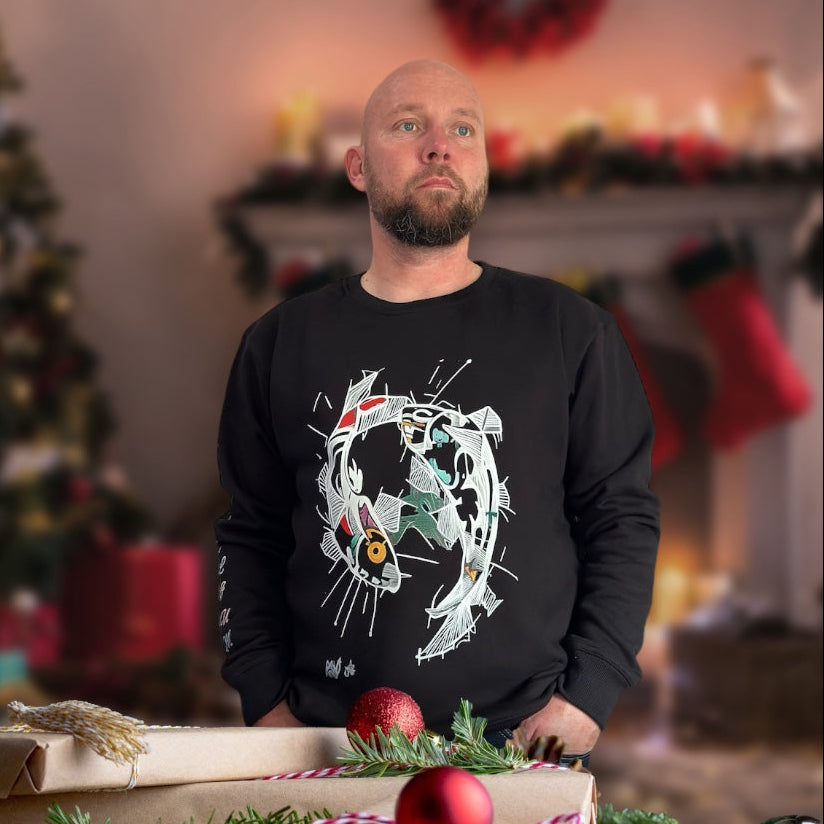Man in a black sweater with a decorative design, standing behind wrapped gifts and Christmas decorations. rokkatei-eshop.mom