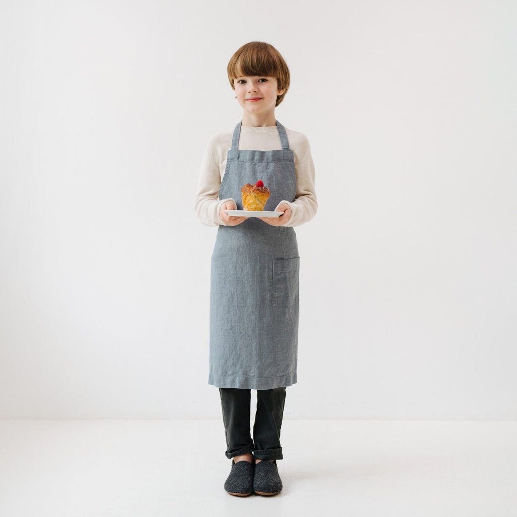 a child's soft blue linen apron with a front pocket, shown laid flat on a wooden table for a practical view of its adjustable ties and durable handmade construction