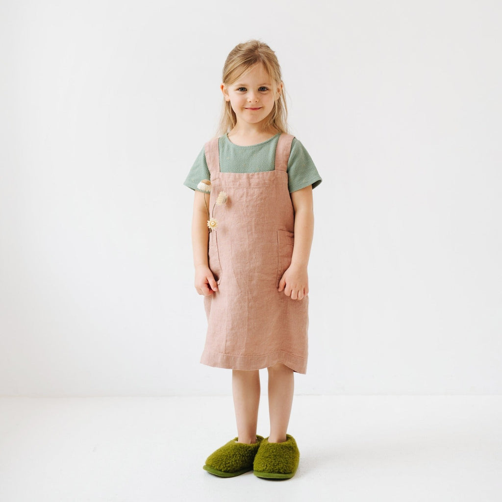 a child laughing while wearing a cream linen pinafore apron, playfully holding a wooden spoon